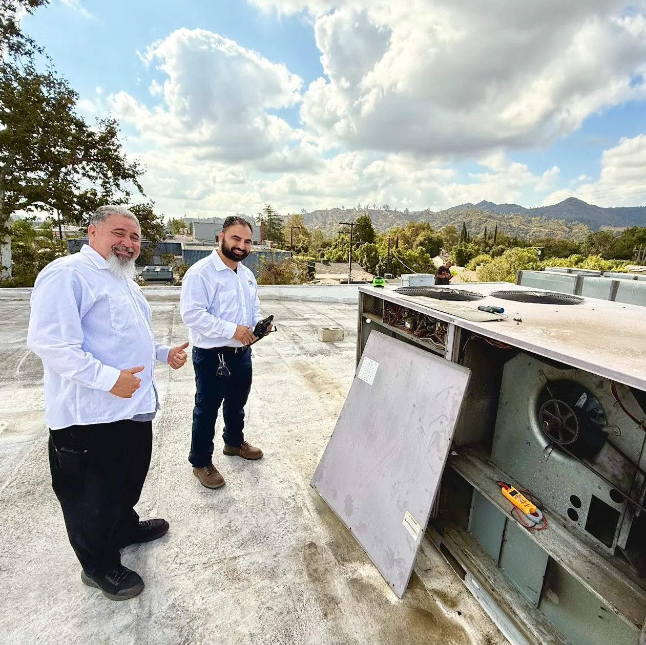 Two people standing beside an open rooftop HVAC unit, with tools on the unit.